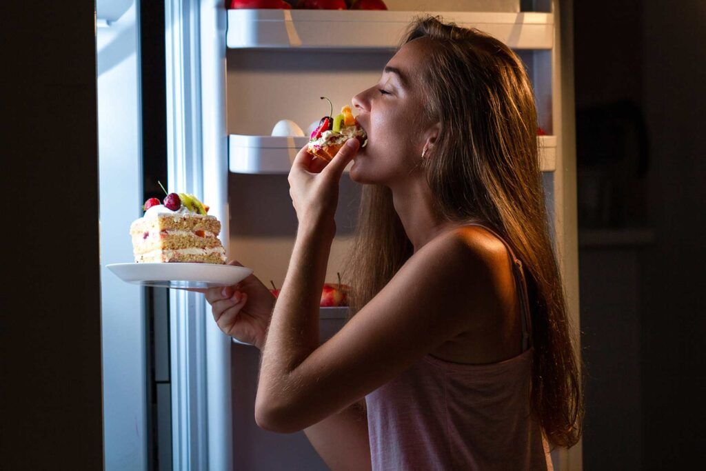 Frau isst bei Dunkelheit Kuchen direkt am Kühlschrank